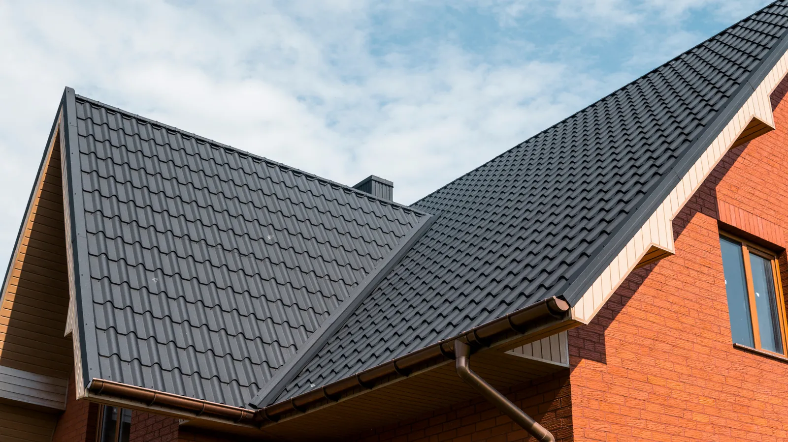 Partly cloudy sky above a brick house with dark metal tile roofing